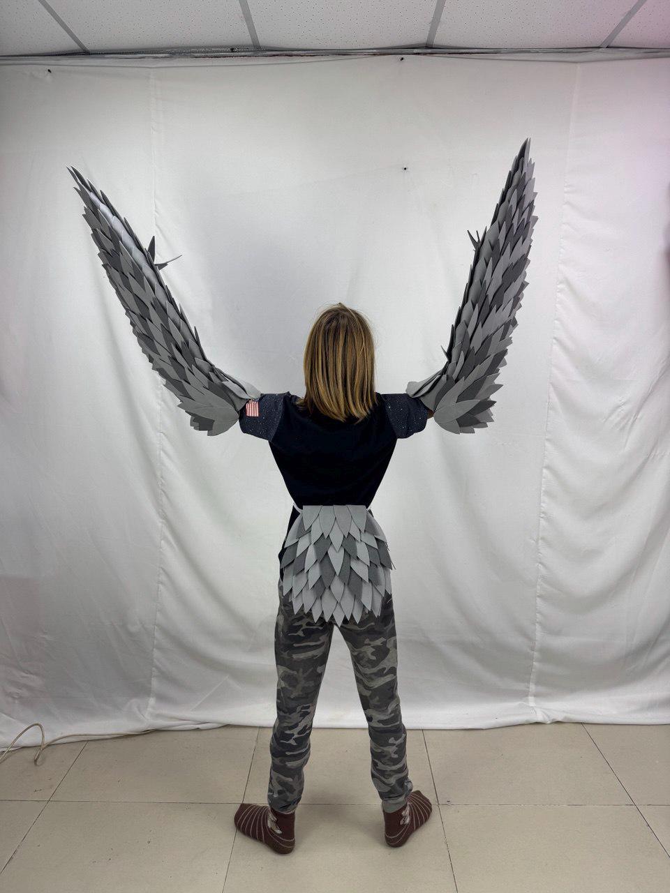 May include: A person wearing a black top and camouflage pants with gray and white bird wings attached to the arms. The wings are made of layered, textured material, and the person is standing with arms outstretched.