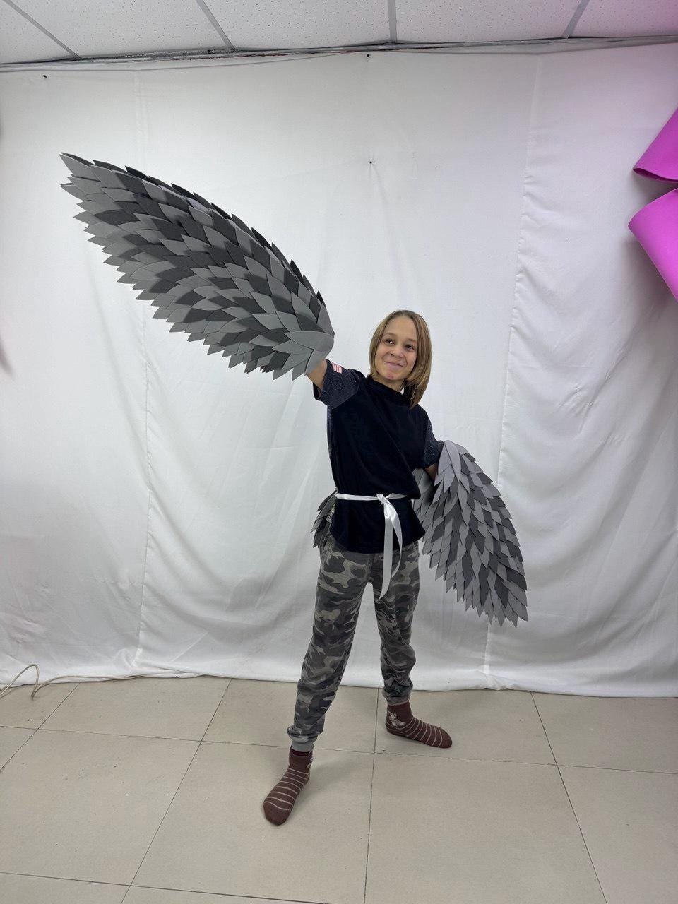 May include: A person wearing a black shirt and camouflage pants with gray and dark gray feathered wings. The wings are attached to the arms and back. The wings are made of layered, diamond-shaped pieces.