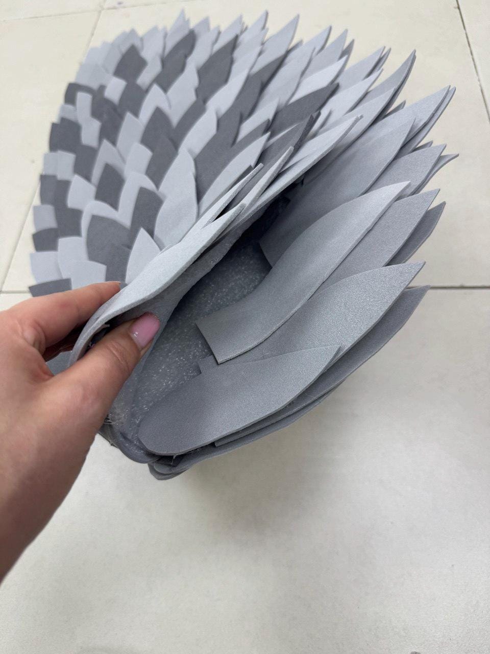 May include: A gray, layered, decorative piece with a fan-like design. The piece is made of felt-like material and features a repeating pattern of leaf-shaped elements in varying shades of gray.