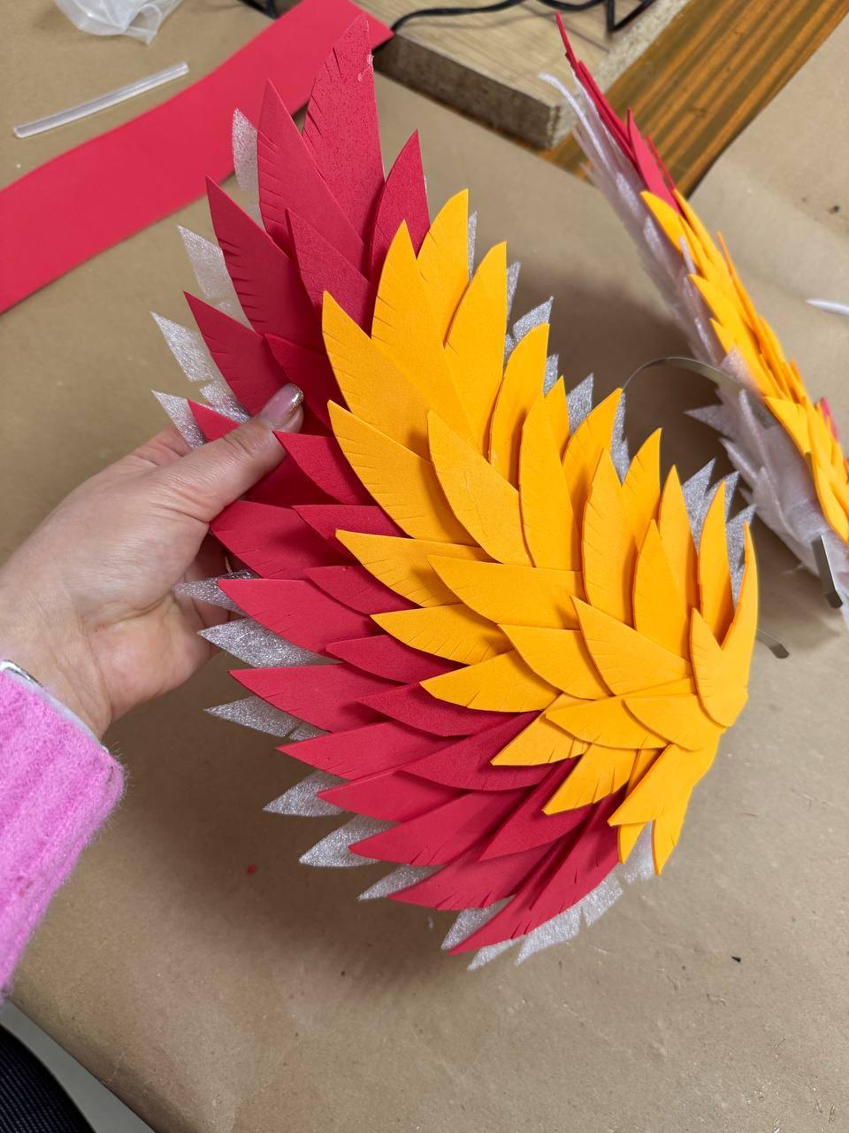 May include: A pair of handmade, layered wings in vibrant red and yellow. The wings are constructed from individual, feather-shaped pieces, creating a textured, layered effect. The wings are likely for a costume or craft project.