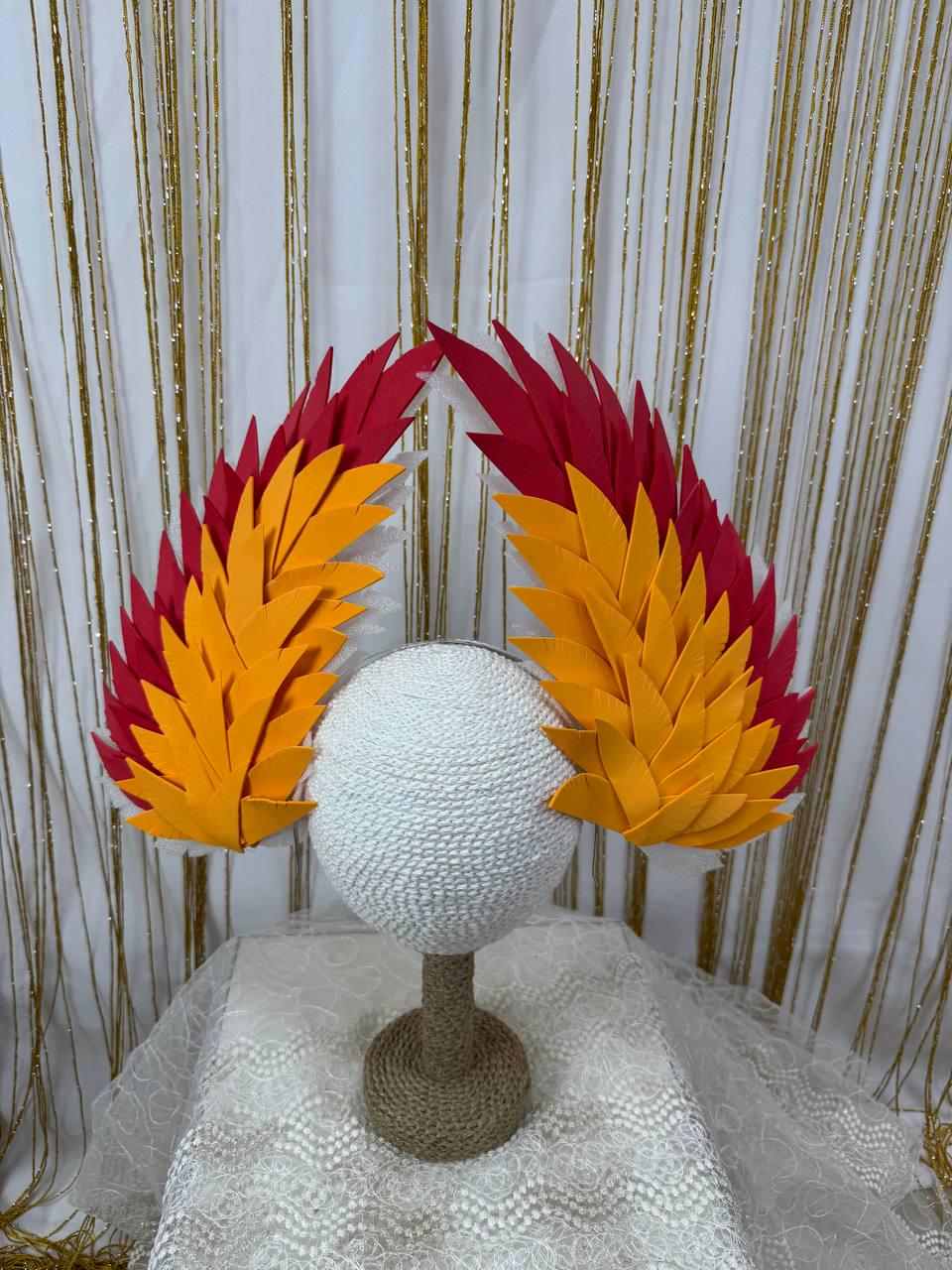 May include: A pair of vibrant, handmade headpieces with red and yellow felt feather designs. The headpieces are displayed on a white, textured sphere atop a brown stand, set against a gold and white backdrop.