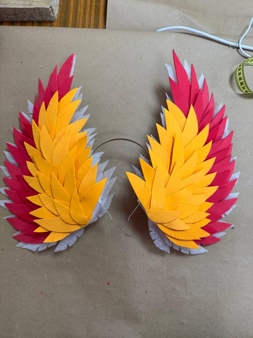 May include: A pair of handmade, colorful costume wings. The wings are made of red, yellow, and orange paper feathers, attached to a silver wire frame. The wings are designed to be worn as a costume accessory.