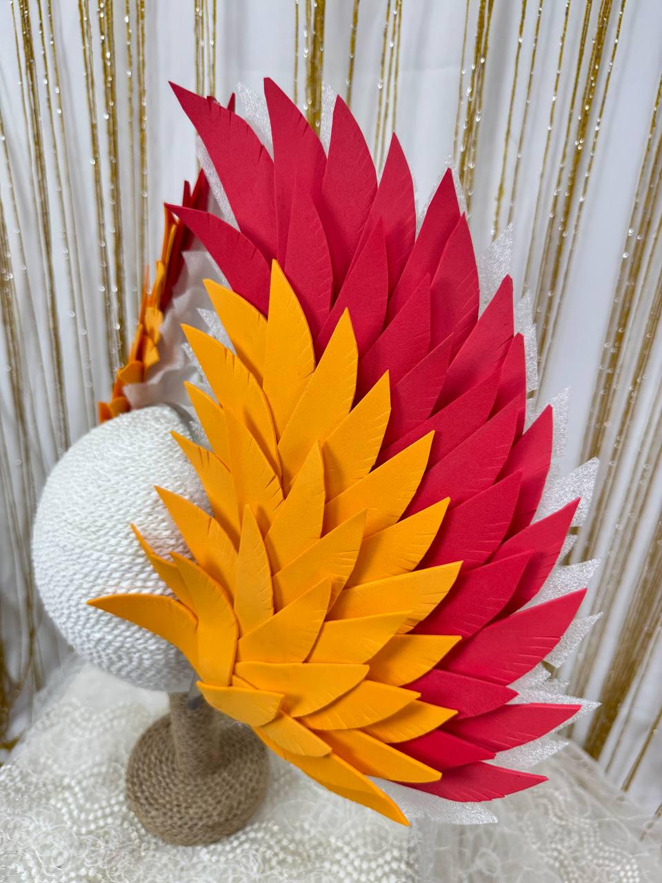 May include: A decorative headpiece featuring red and yellow feather-shaped elements. The headpiece is mounted on a white sphere and a brown base. The background includes a white curtain and gold string lights.