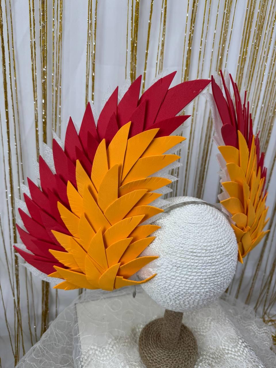 May include: A vibrant headpiece featuring red and orange feather-like elements. The design is mounted on a white, spherical base, suggesting a costume or theatrical accessory. The feathers are arranged in a layered pattern.