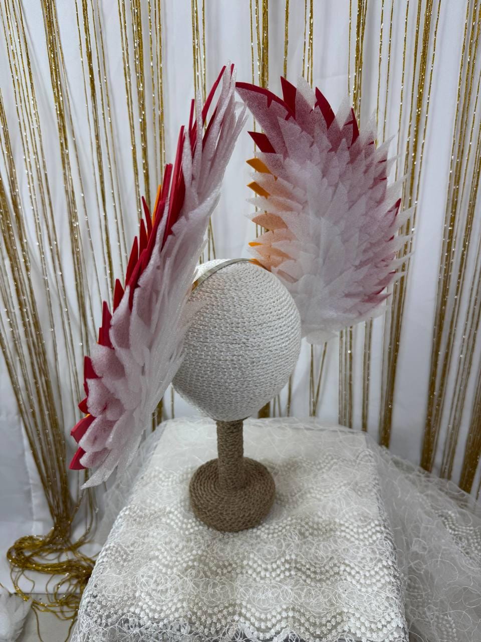 May include: A decorative headpiece with white, red, and orange accents, displayed on a mannequin head. The headpiece features layered, feather-like elements, creating a dramatic, artistic design. The base is a white, woven material, and the stand is wrapped in twine.