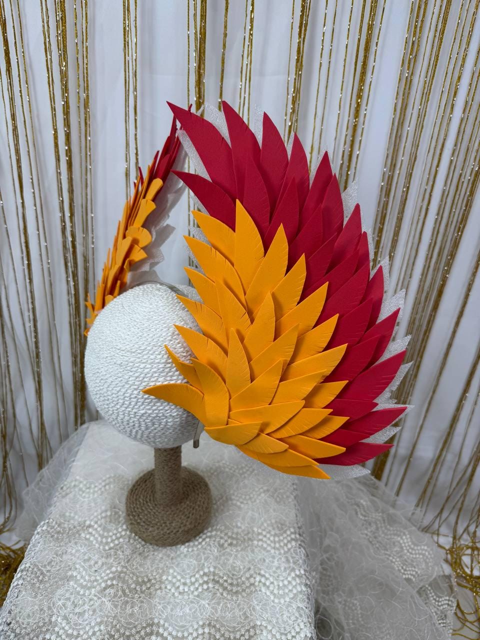 May include: A decorative headpiece with red and yellow feather-like accents. The headpiece is mounted on a white sphere and a brown stand. The background features a white curtain and gold string lights.