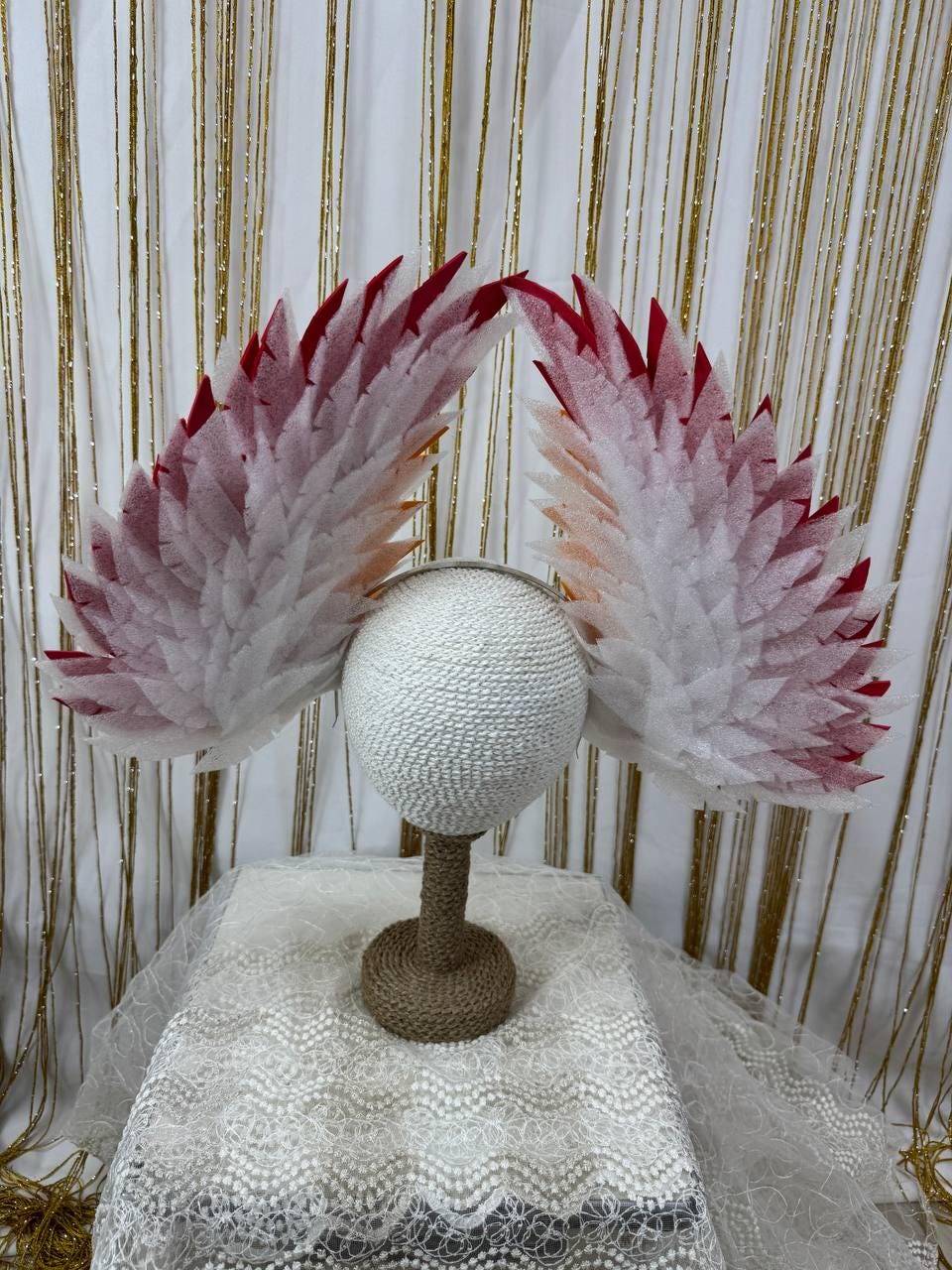 May include: A white headpiece with large, feathery wings. The wings are white with red tips, and the headpiece is on a white, textured stand. The background is a gold and white curtain.