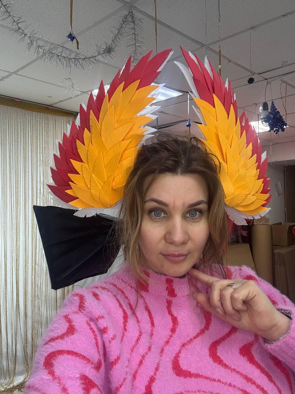 May include: A woman wearing a pink sweater and a headpiece with large, colorful wings. The wings feature red, yellow, and white layered feathers. The background includes a white ceiling and decorative elements.