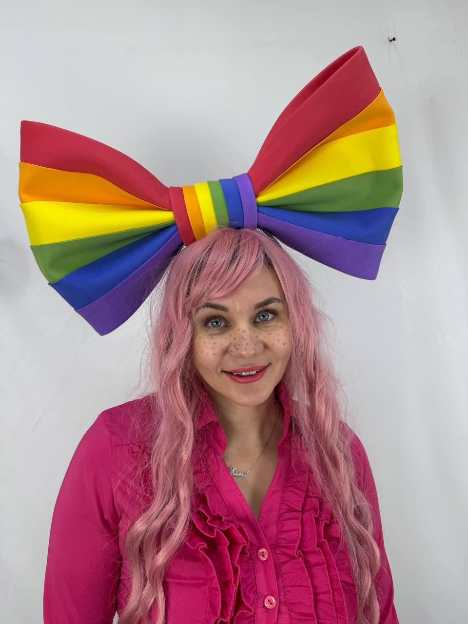 May include: A large, colorful rainbow bow, featuring red, orange, yellow, green, blue, and purple stripes. The bow is worn on the head. The person is wearing a pink shirt.
