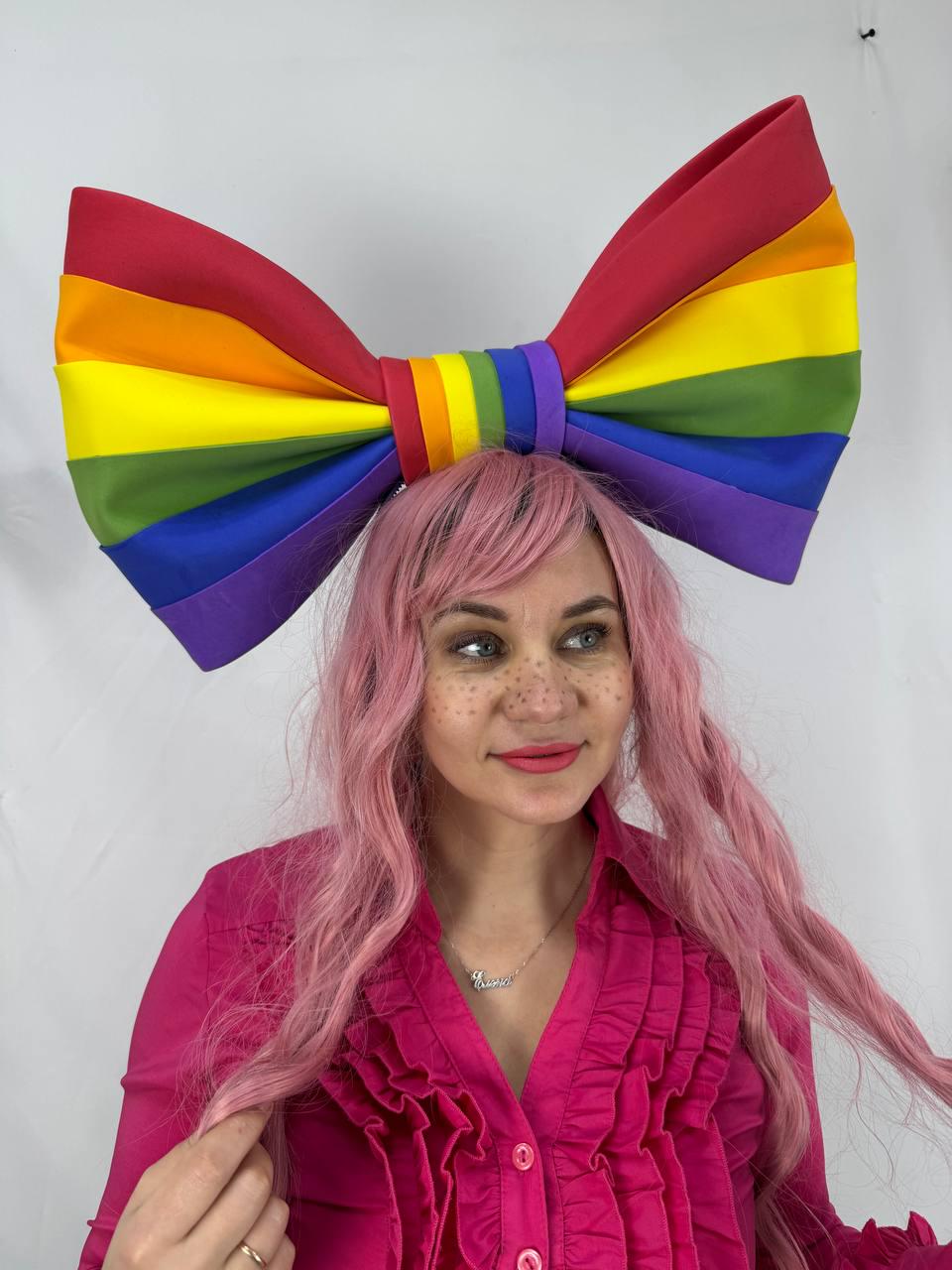 May include: A large, rainbow-colored bow headpiece. The bow features the colors of the rainbow: red, orange, yellow, green, blue, and purple. The headpiece is worn by a person wearing a pink shirt.