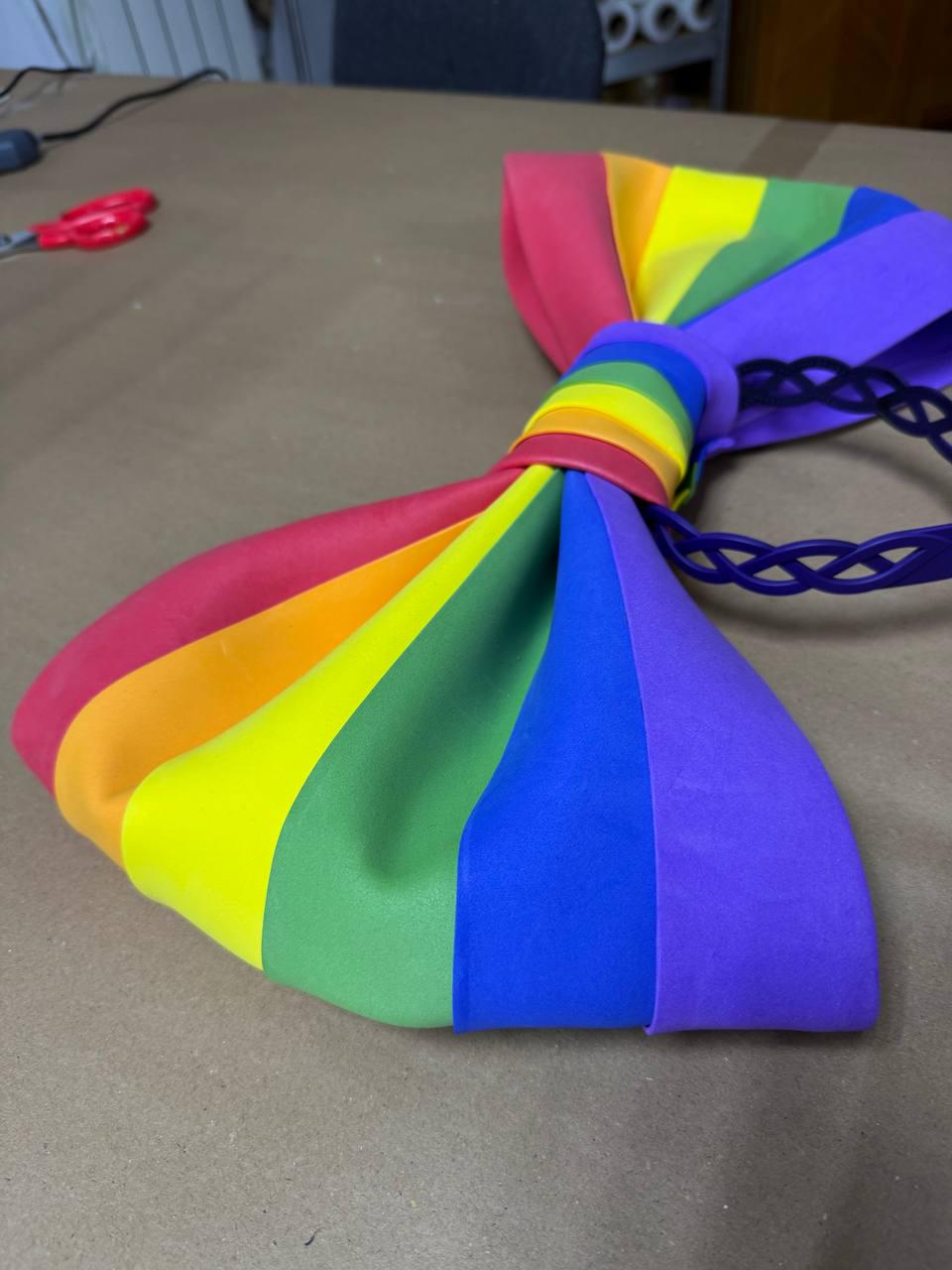 May include: A large, colorful bow tie in a rainbow pattern. The bow tie features the colors of the rainbow, including red, orange, yellow, green, blue, and purple. It has a purple headband attached.