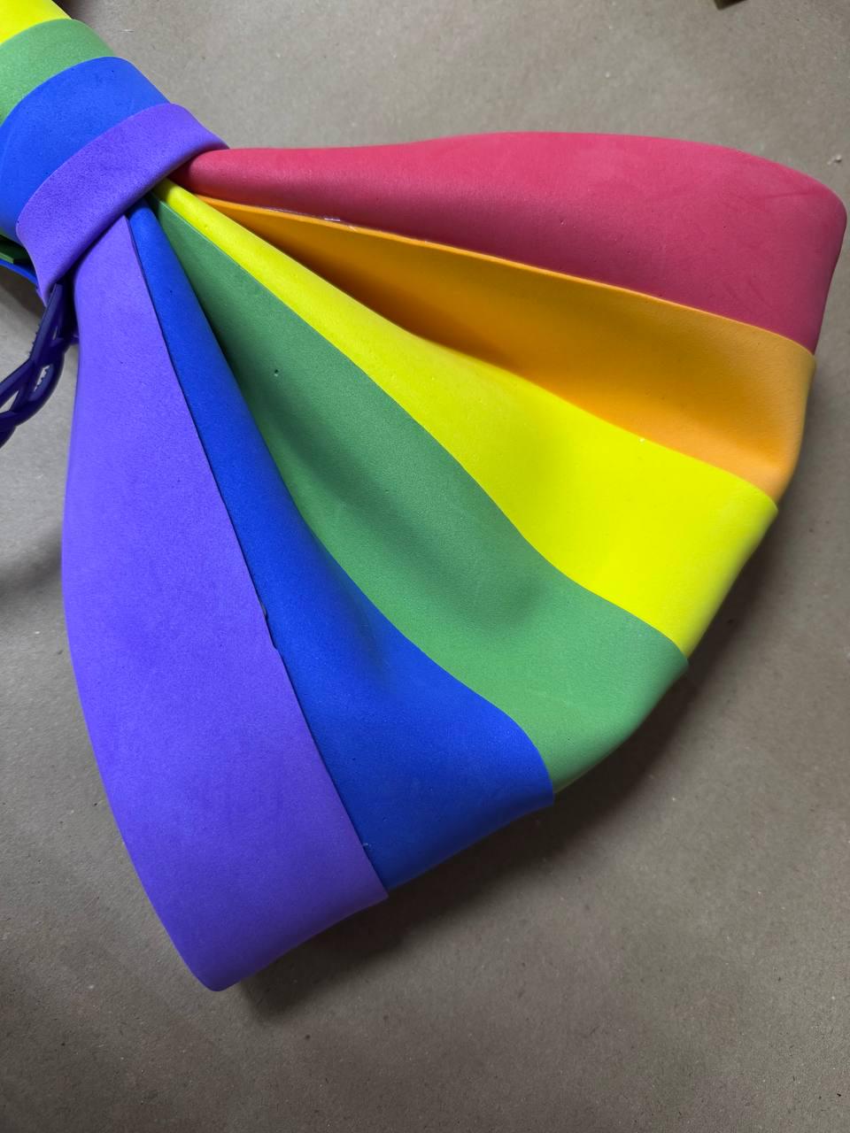 May include: A colorful rainbow-striped bow tie. The bow tie features the colors of the rainbow: red, orange, yellow, green, blue, and purple. The bow tie is made of a soft material and is tied with a dark purple string.