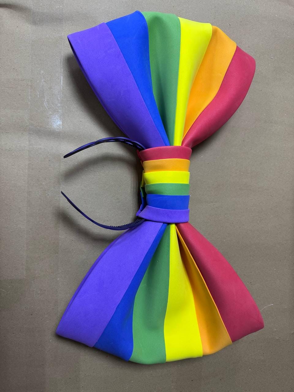 May include: A large, colorful bow tie in a rainbow spectrum of colors: purple, blue, green, yellow, orange, and red. The bow tie is made of a smooth material and has a dark blue adjustable neckband.