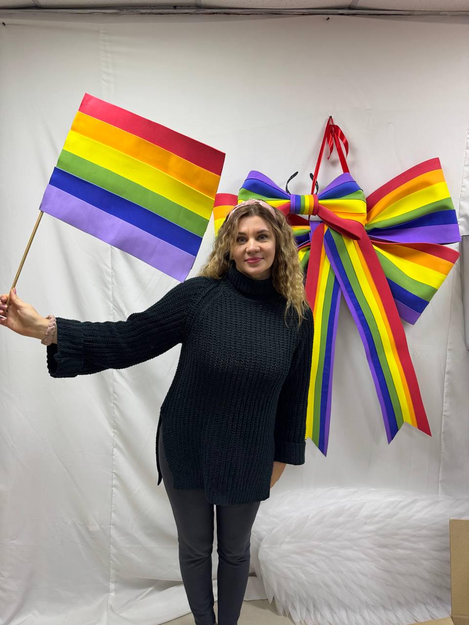 May include: A woman holding a rainbow flag, with a large rainbow bow decoration hanging on the wall. The flag and bow feature the colors of the LGBTQ+ pride flag. The woman is wearing a black sweater and gray pants.