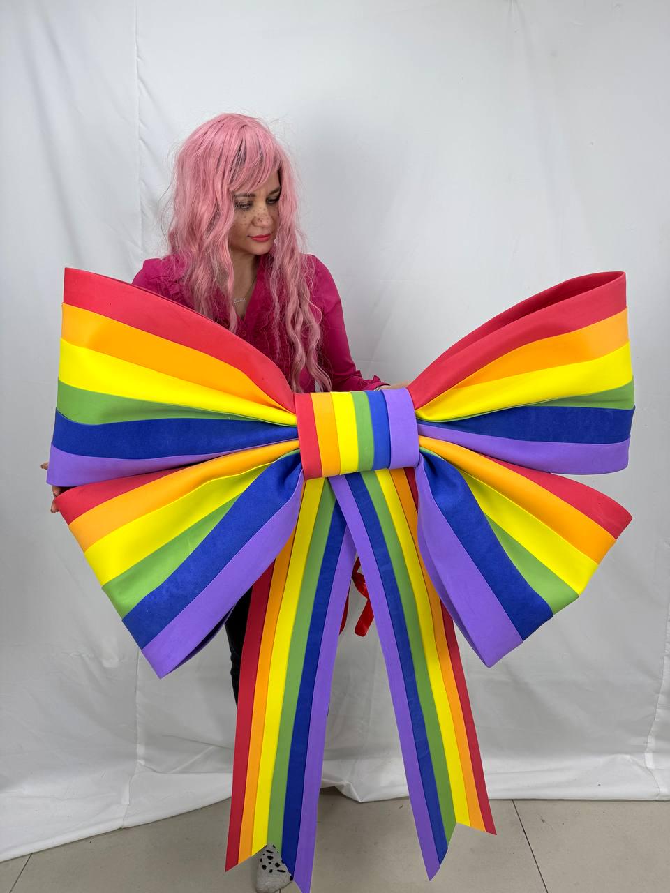May include: A large, colorful rainbow bow with red, orange, yellow, green, blue, and purple stripes. The bow is held up, showcasing its vibrant colors and design. The bow's size suggests it could be used for decoration or as a prop.