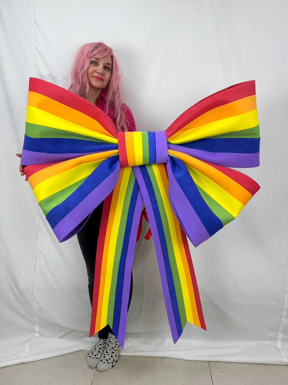 May include: A large, colorful rainbow bow with red, orange, yellow, green, blue, and purple stripes. The bow is a vibrant decoration, perfect for celebrations or events. The bow's dimensions are approximately 48 inches wide.