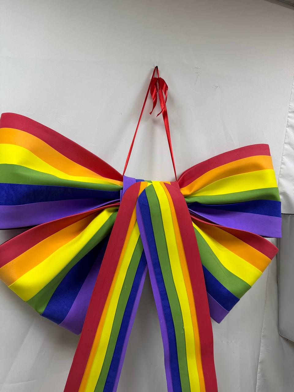 May include: A large, decorative bow in the colors of the rainbow, including red, orange, yellow, green, blue, and purple. The bow is made of a satin-like material and has long, vertical streamers. A red ribbon is used to hang the bow.