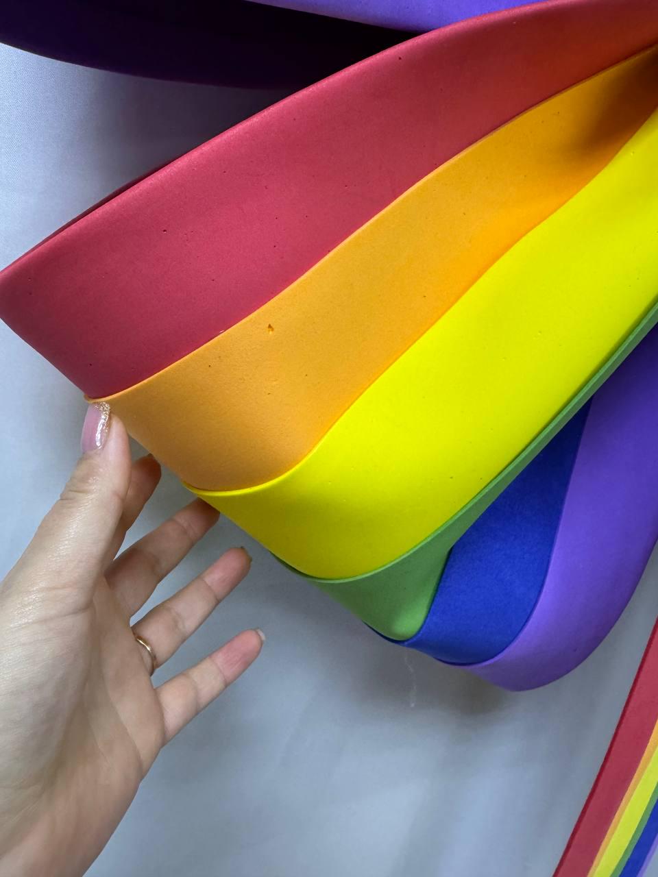 May include: A vibrant rainbow-colored object with red, orange, yellow, green, blue, and purple bands. The object is curved and appears to be made of a soft material. The image is a close-up, highlighting the bright colors.