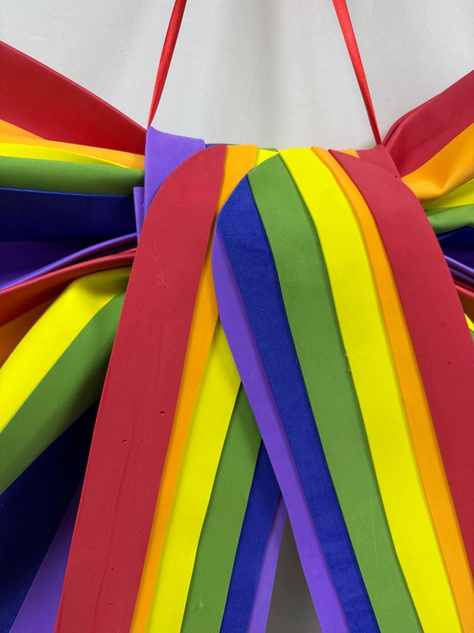 May include: A colorful rainbow-themed decoration with a red ribbon for hanging. The design features layered, curved strips in the colors of the rainbow: red, orange, yellow, green, blue, and purple. The decoration is made of a soft, flexible material.