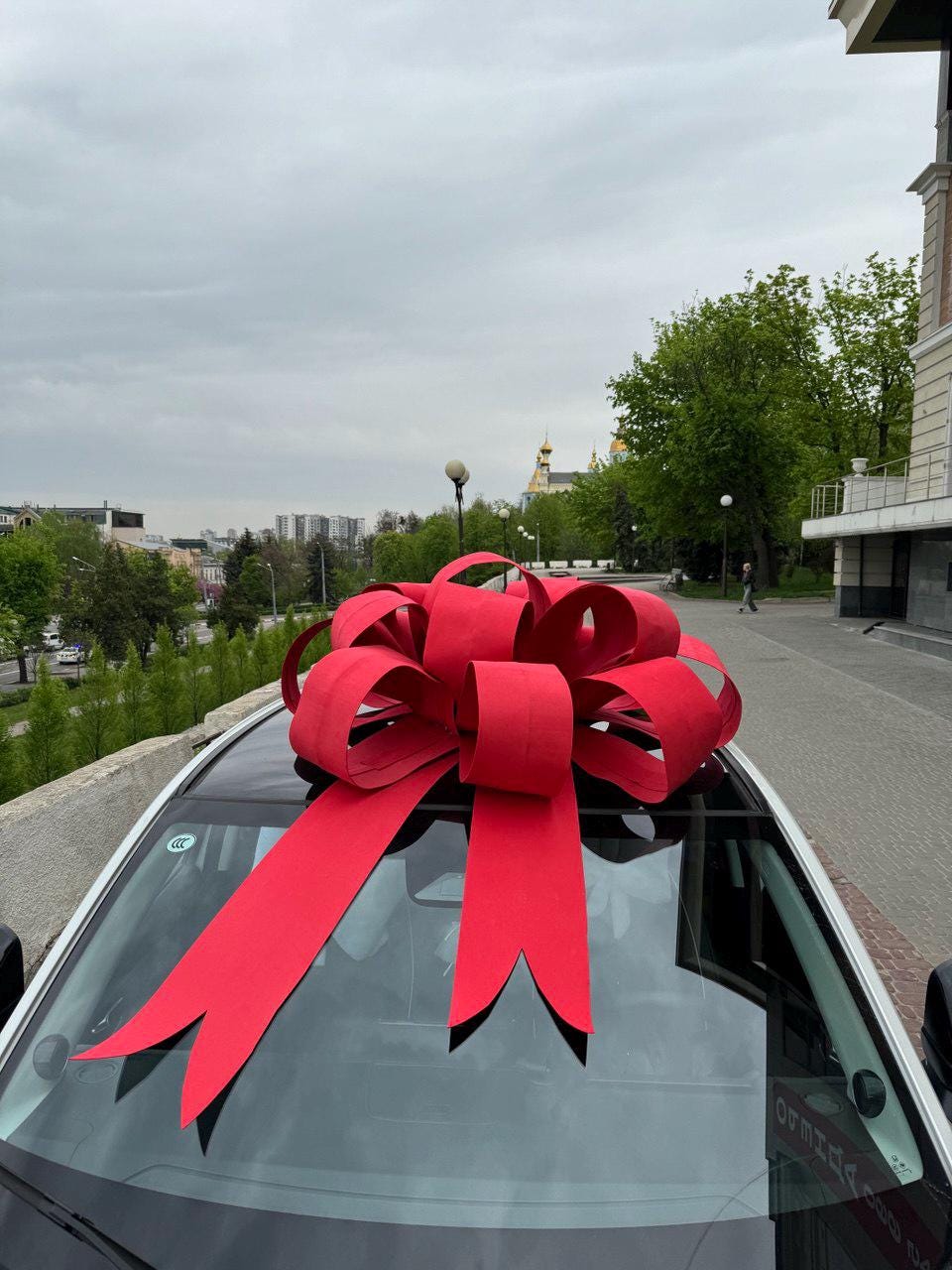 May include: A large, bright red bow adorns the roof of a car, suggesting a celebratory occasion. The bow's ribbons cascade down the windshield. The car is parked outdoors, with a cityscape and overcast sky in the background.