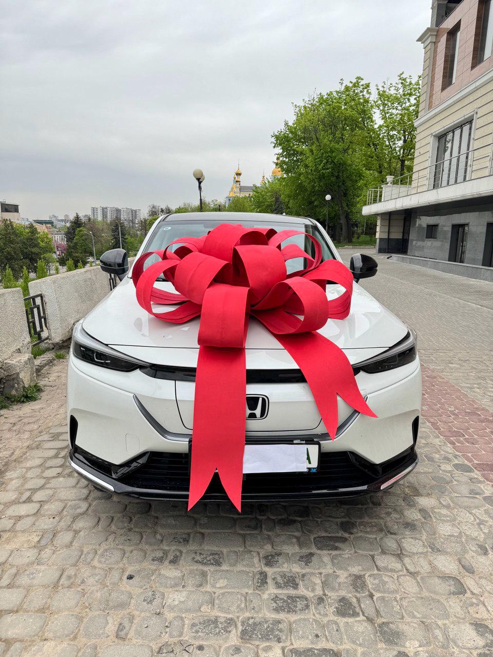 May include: A white Honda SUV is adorned with a large, vibrant red bow, suggesting a gift or celebration. The car is parked on a cobblestone surface, with a backdrop of buildings and greenery under an overcast sky.