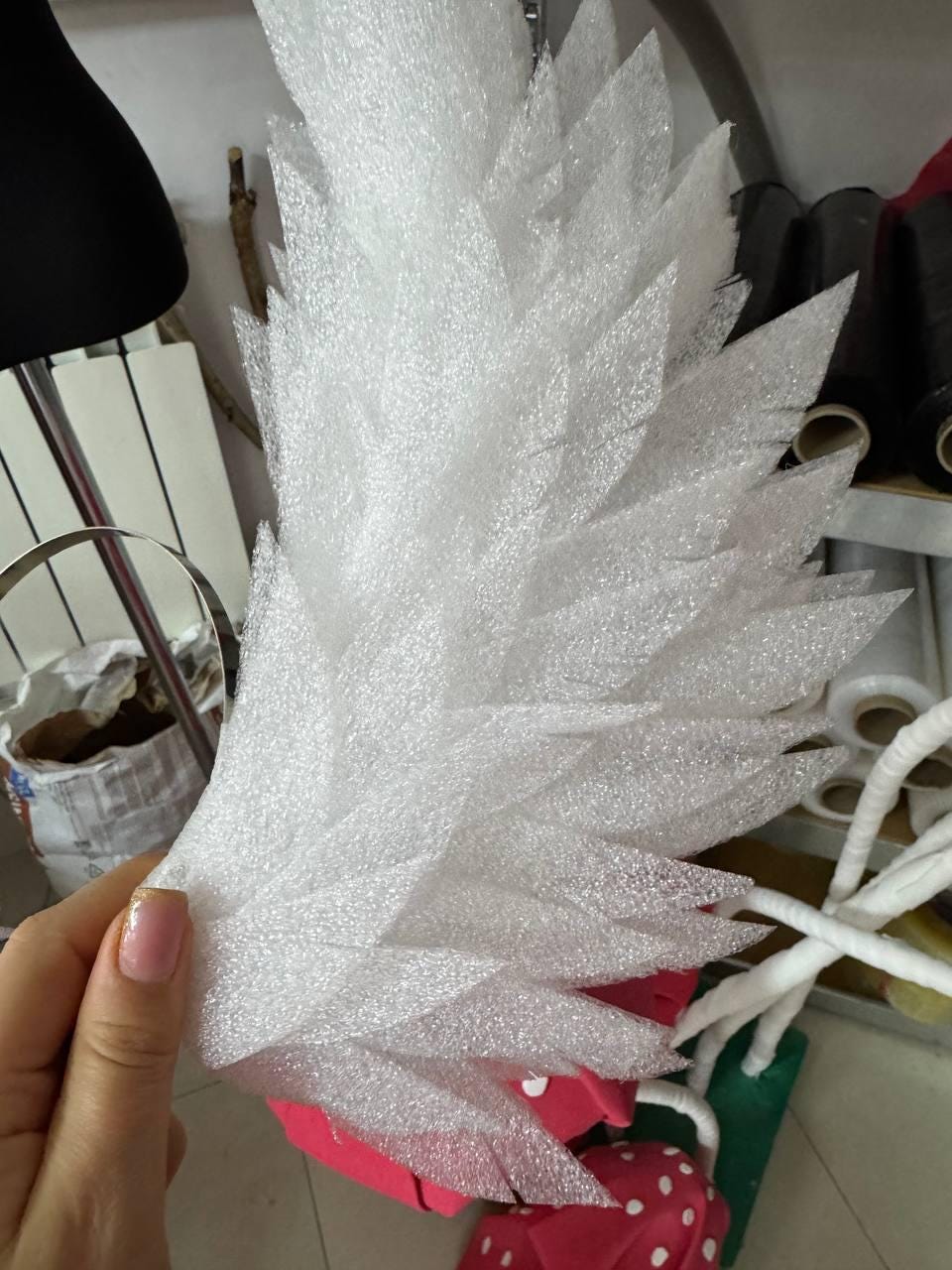 May include: A large, white, feather-shaped craft element made of textured foam. The foam is cut into individual, pointed shapes, resembling feathers. The craft item is held in a hand, with other craft supplies visible in the background.
