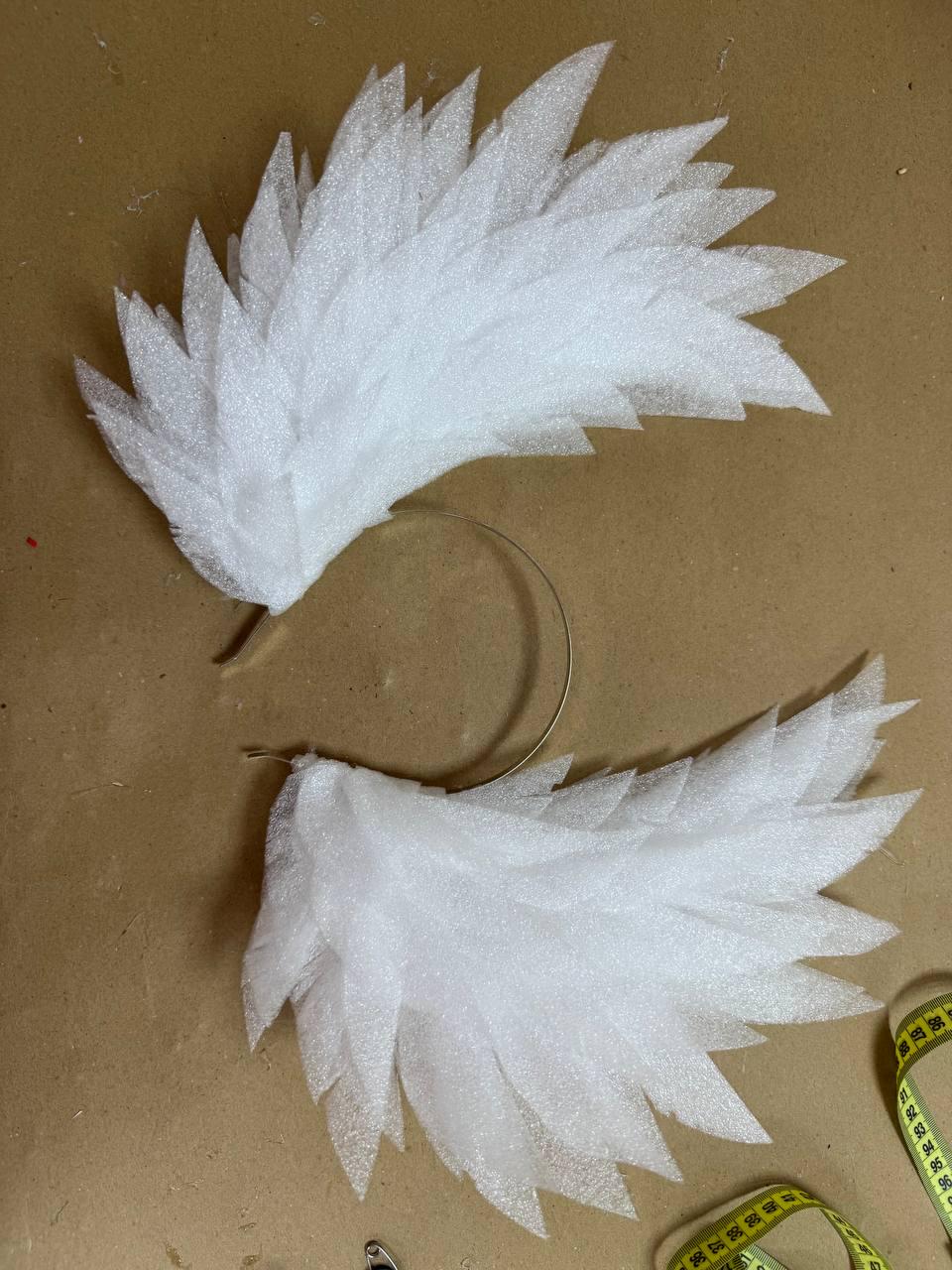 May include: Two white, feathery angel wing headbands. Each wing is made of layered, textured foam and attached to a thin, gold-colored wire headband. The wings are designed to be worn on the head.