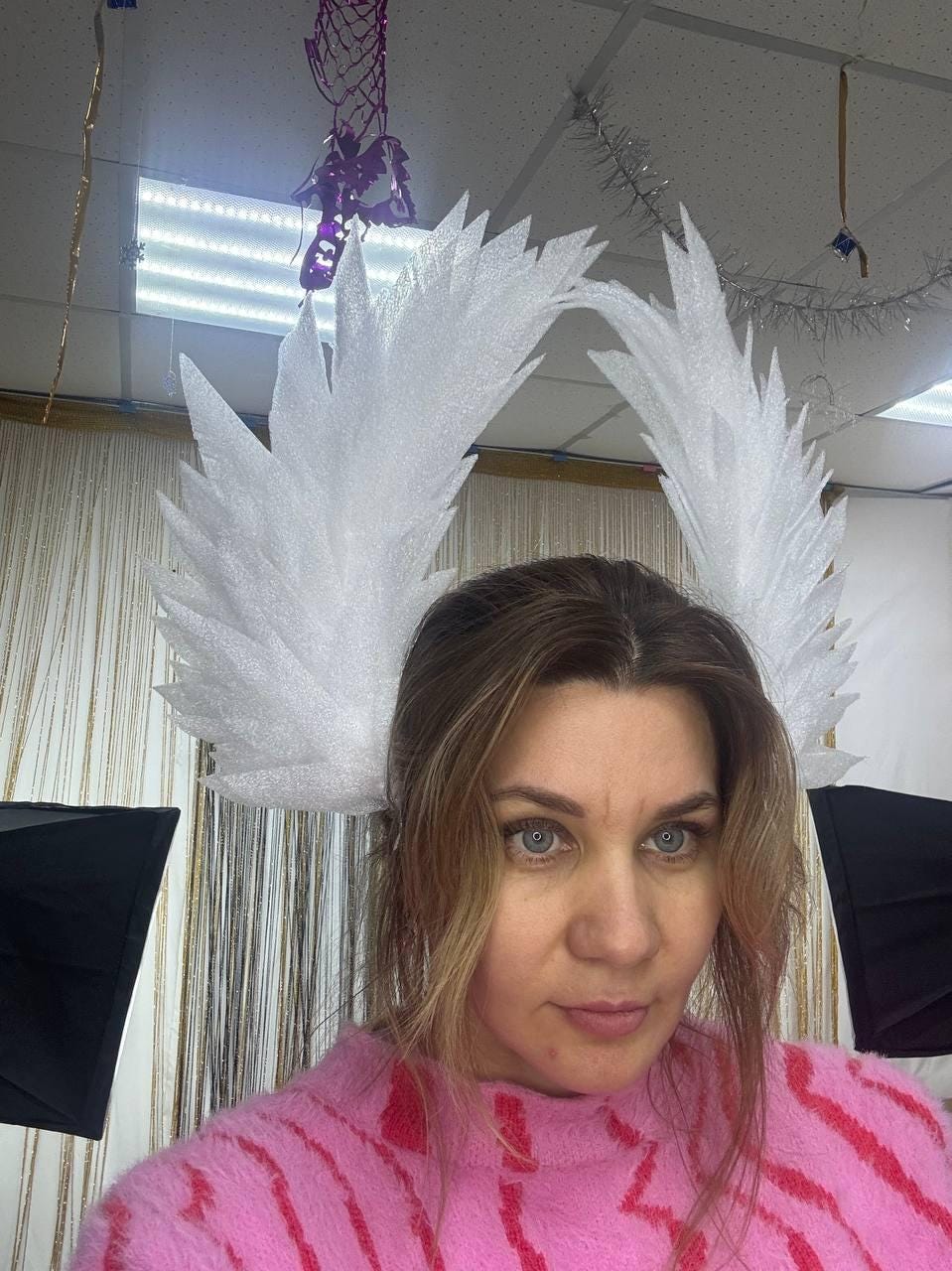 May include: A white, feathery headpiece with a pointed design. The headpiece is worn on the head. The person in the image is wearing a pink sweater with red stripes.