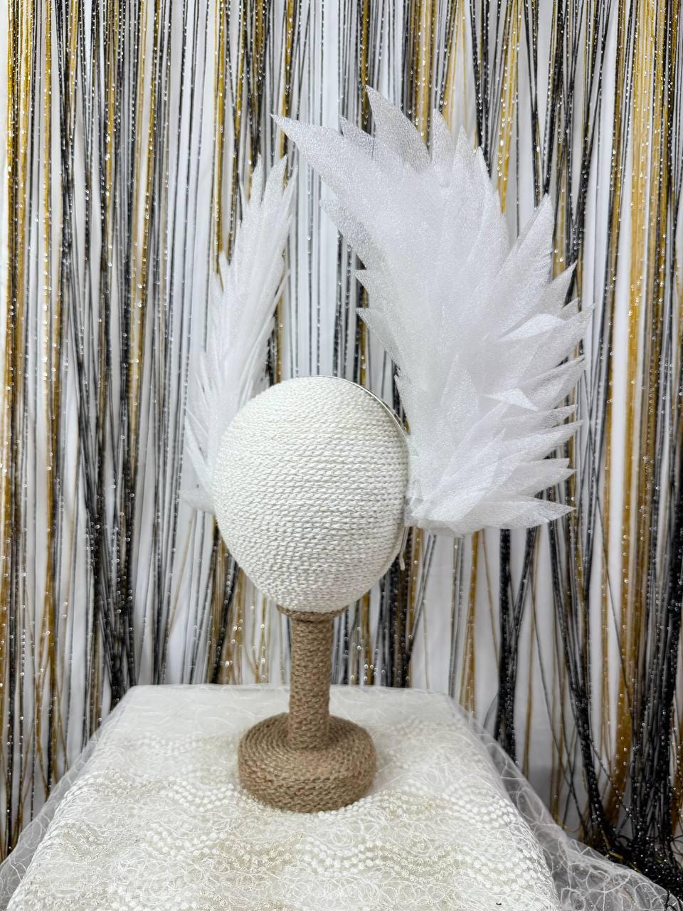 May include: A white, textured head form with attached white feather-like accents, displayed on a jute-wrapped stand. The stand sits on a white lace-covered surface, with a backdrop of gold, black, and white shimmering strands.