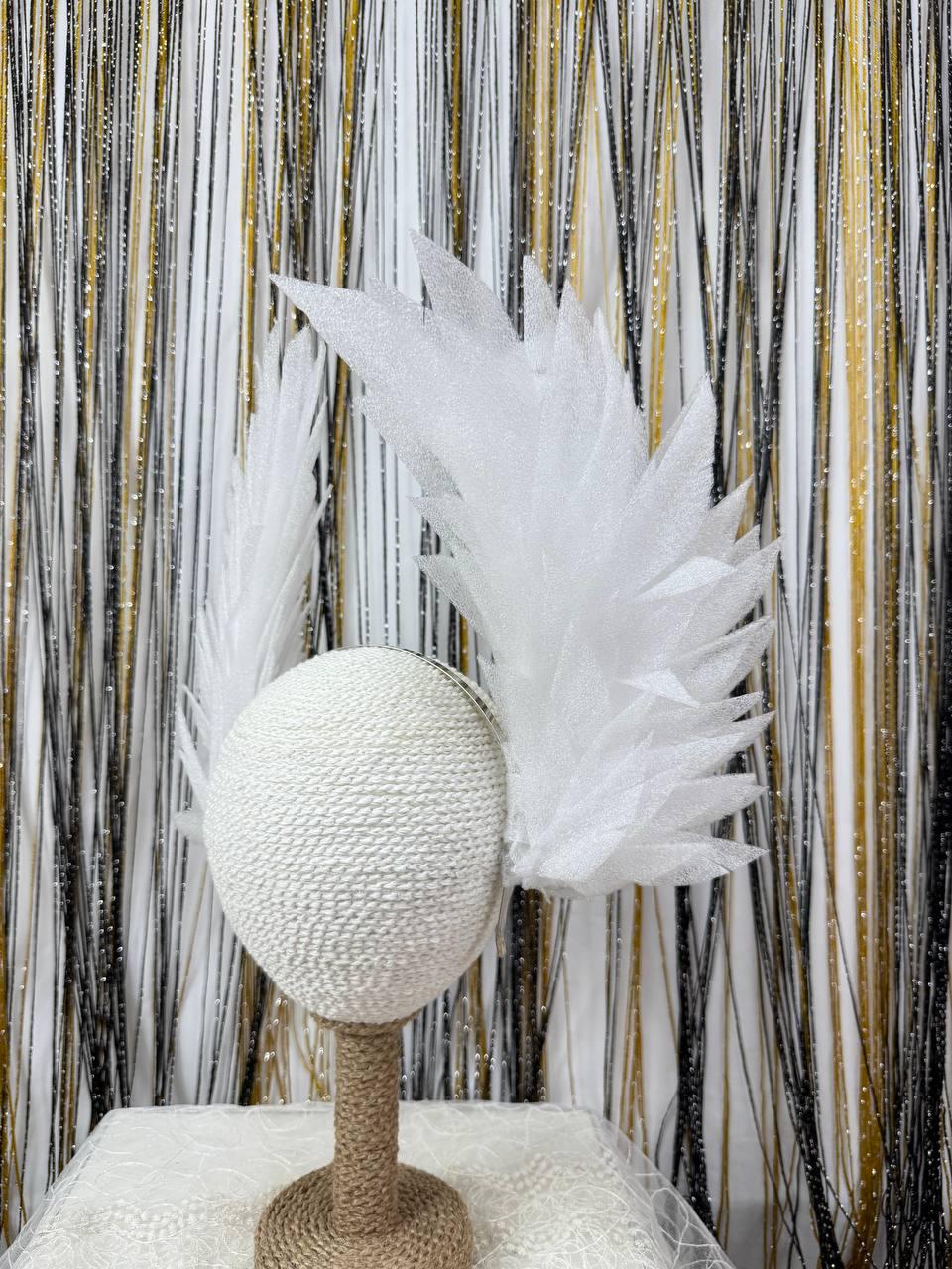 May include: A white, textured headpiece with feathery, wing-like accents. The headpiece is mounted on a decorative stand. The background features a curtain of black, gold, and white strands.