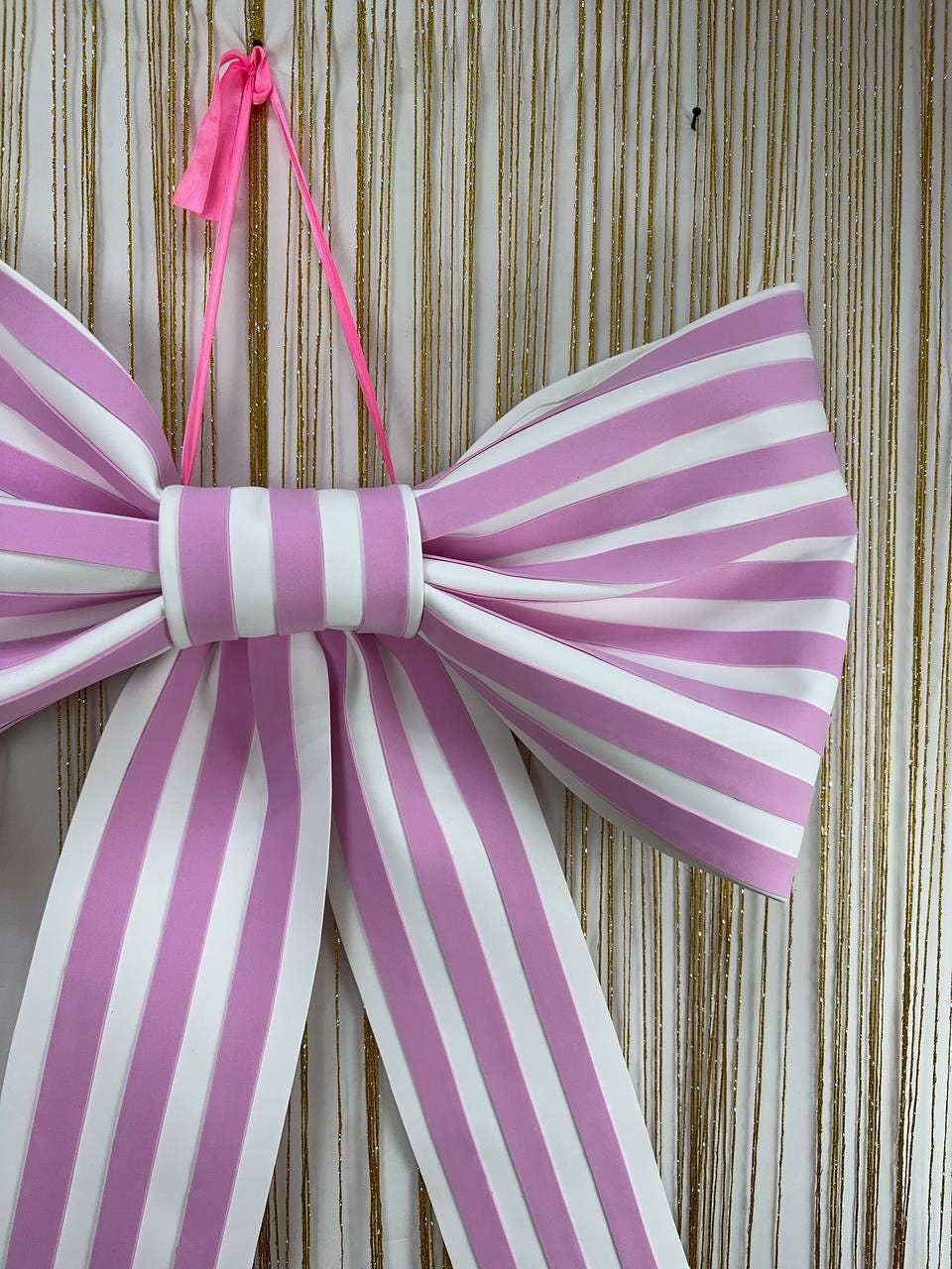 May include: A large, decorative bow with alternating pink and white stripes. The bow is suspended by a pink ribbon. The bow's design features a central knot and long, flowing tails, perfect for party decorations.