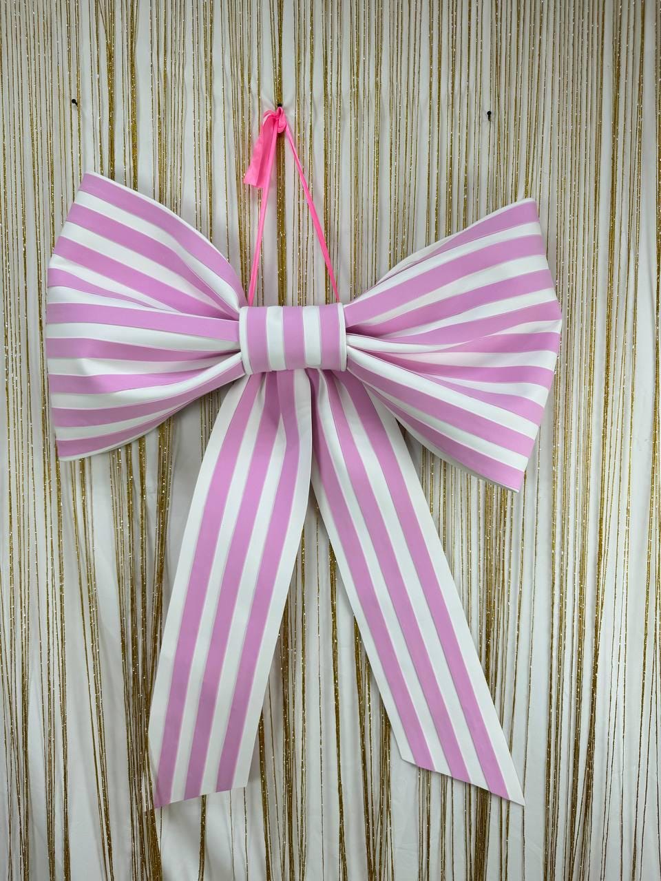 May include: A large, decorative bow with pink and white vertical stripes. The bow is suspended by a pink ribbon and is against a gold and white striped background. The bow's design features a central knot and long, flowing tails.