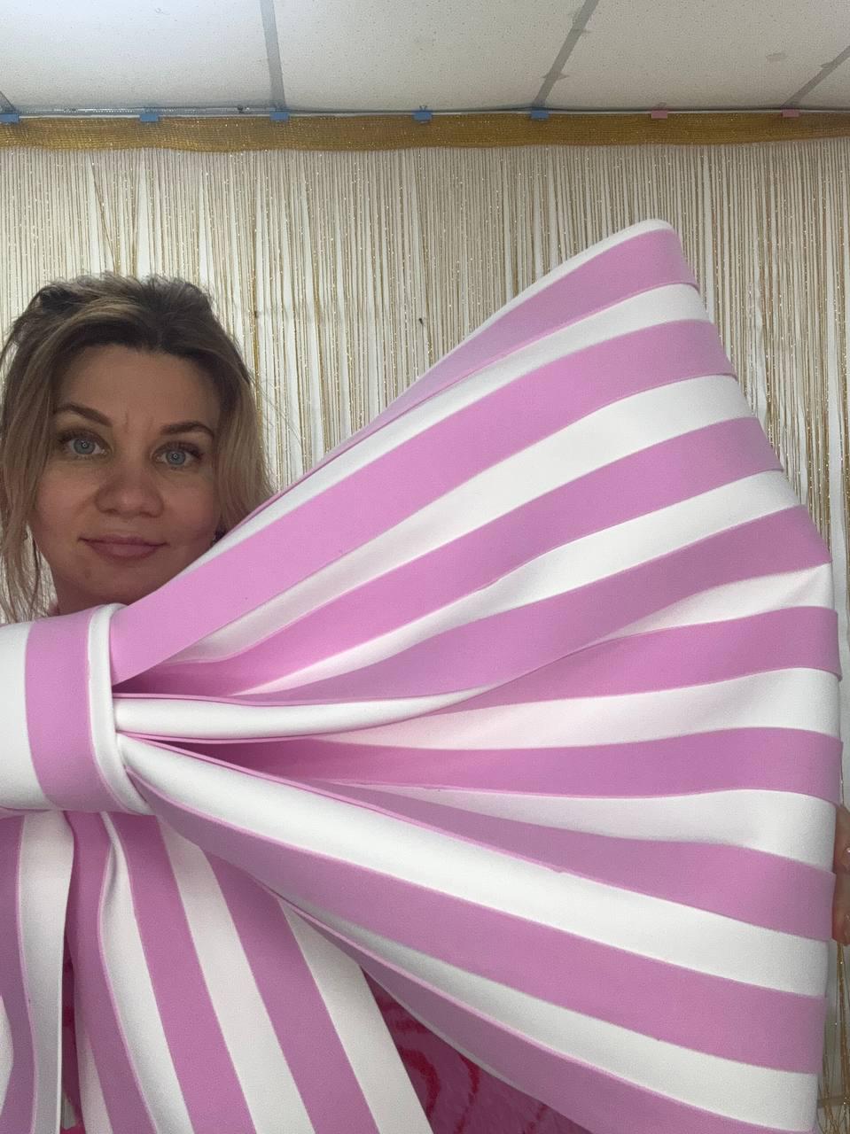 May include: A large, decorative bow with alternating pink and white stripes. The bow is held up, showcasing its size and design. The stripes are evenly spaced, creating a bold visual statement. The bow's knot is centered, adding to its symmetrical appeal.