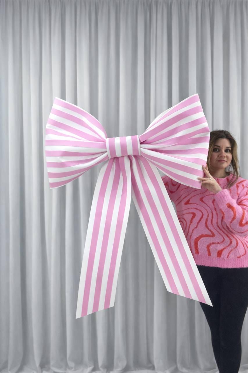 May include: Large pink and white striped bow. The bow is a decorative element, possibly for a party or event. The bow is held up in front of a white curtain.