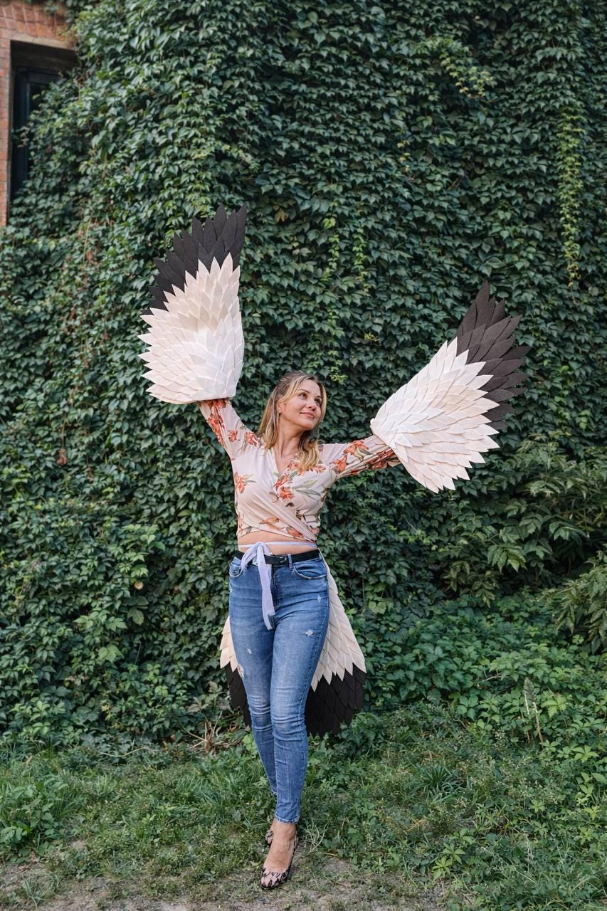 May include: A person wearing large, handmade bird wings with white, beige, and black feathers. The wings are spread wide against a backdrop of green foliage. The person is wearing a floral top and blue jeans.