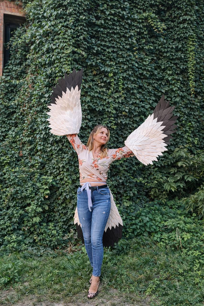 May include: A person wearing large, handmade bird wings with white, beige, and black feathers. The wings are spread wide against a backdrop of green foliage. The person is wearing a floral top and blue jeans.