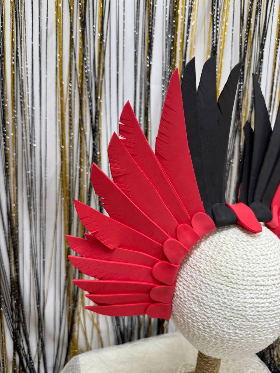 May include: A decorative headpiece featuring red and black feather-like elements. The headpiece is mounted on a white, spherical base wrapped in textured twine. The background includes gold and silver tinsel strands.