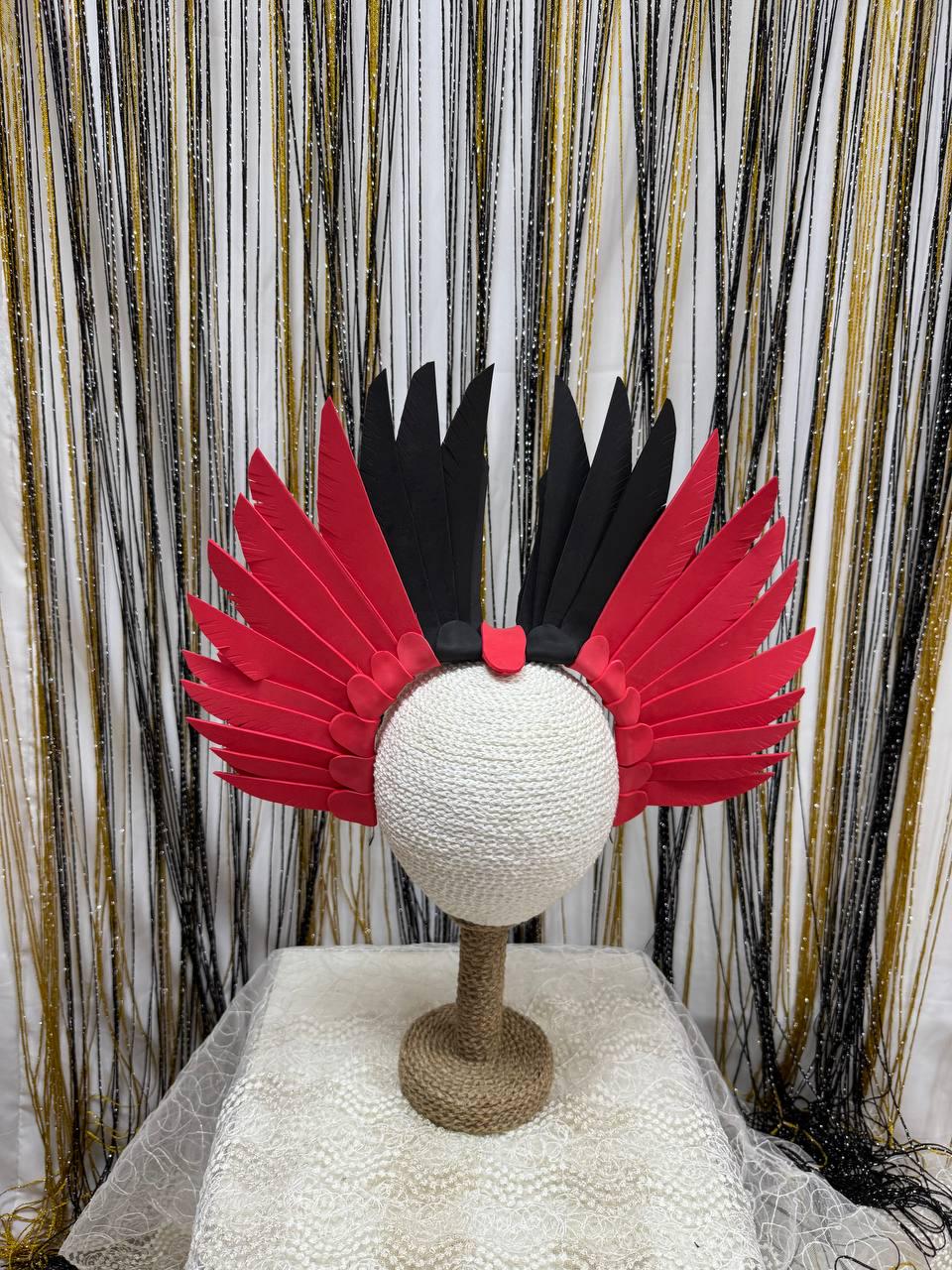May include: A decorative headpiece with red and black feather-like accents. The headpiece is displayed on a white mannequin head. The feathers are arranged in a symmetrical pattern, with red feathers on the sides and black feathers in the center.