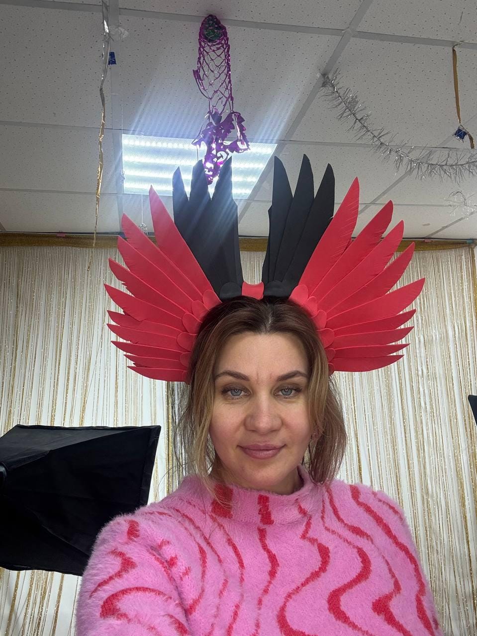 May include: A woman wearing a pink sweater and a decorative headpiece. The headpiece features red and black feather-like elements. The sweater has a pink and red pattern.