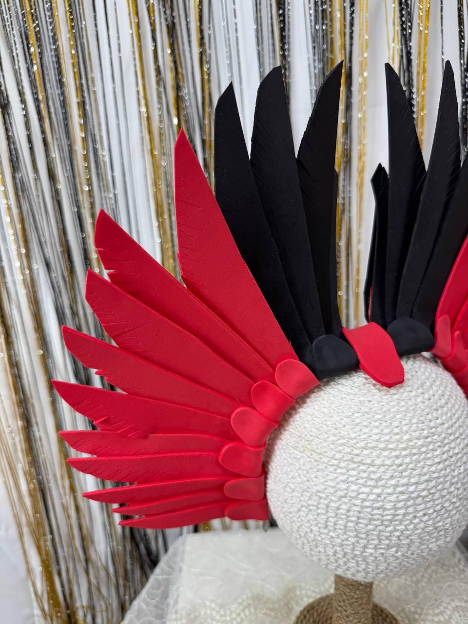 May include: A decorative headpiece featuring red and black feather-like elements. The headpiece is attached to a white, spherical base with a textured, woven appearance. The feathers are arranged in a fan-like pattern.