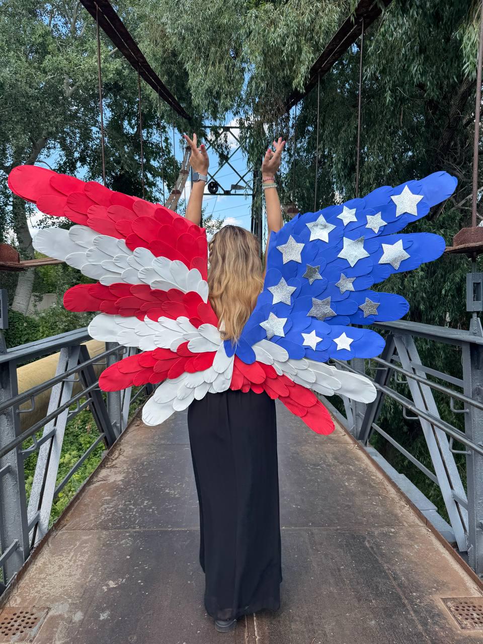 May include: A person wearing patriotic-themed wings, with red, white, and blue feathers. The wings are designed to resemble the American flag, with stars and stripes. The person is standing on a bridge, with arms raised.