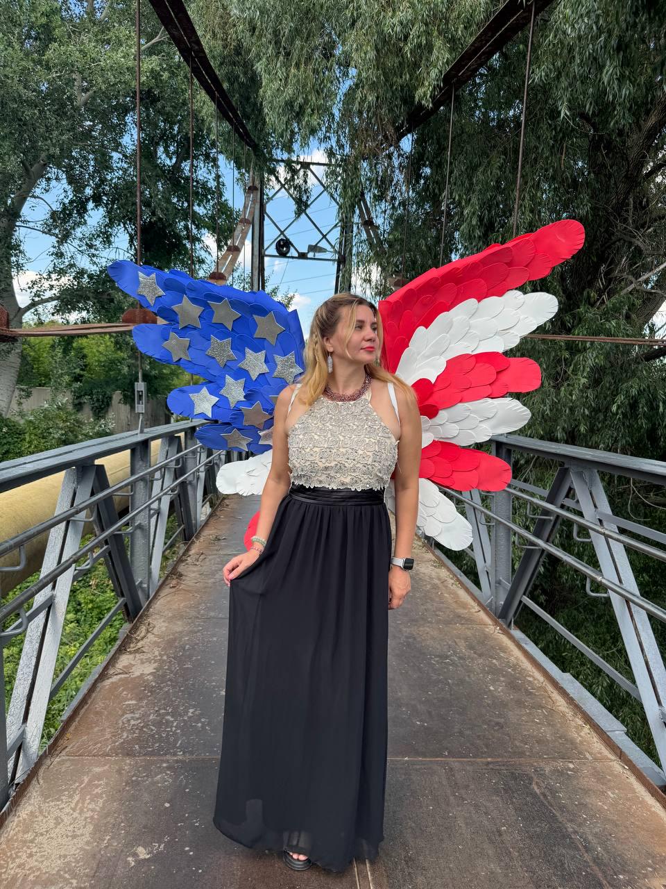 May include: A woman wearing a white and silver beaded top and a black maxi skirt stands on a bridge. Behind her are large decorative wings in the colors of the American flag: red, white, and blue with silver stars.