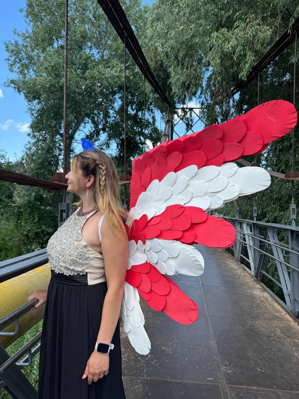 May include: A person wearing large red, white, and blue decorative wings, standing on a bridge. The wings are layered with a feather-like design. The person is wearing a black dress and a silver top.