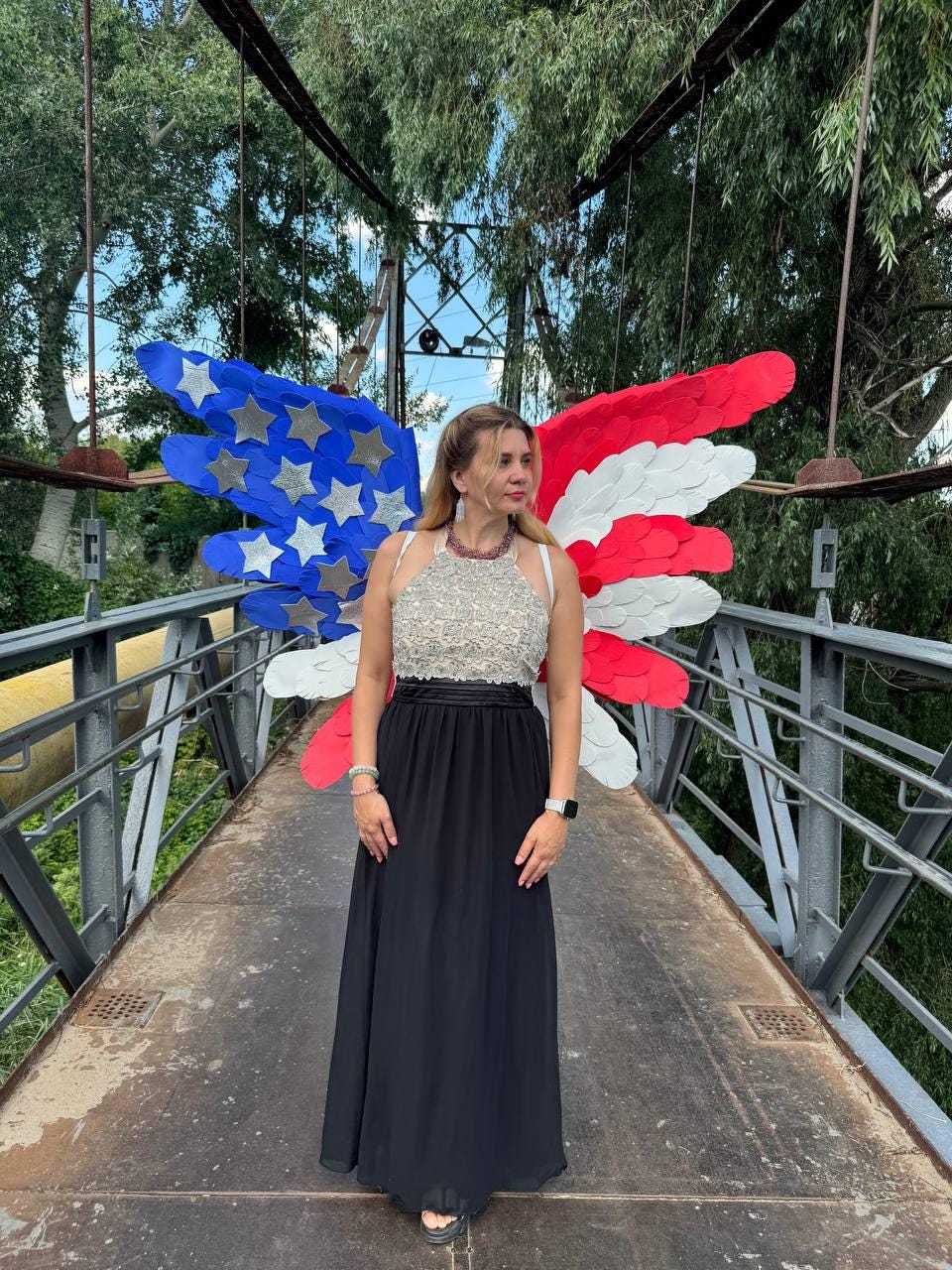 May include: A woman wearing a formal black gown with a beaded top stands on a bridge. Behind her are large, decorative wings in the colors of the American flag: blue with silver stars, and red and white stripes. The scene is outdoors with trees in the background.