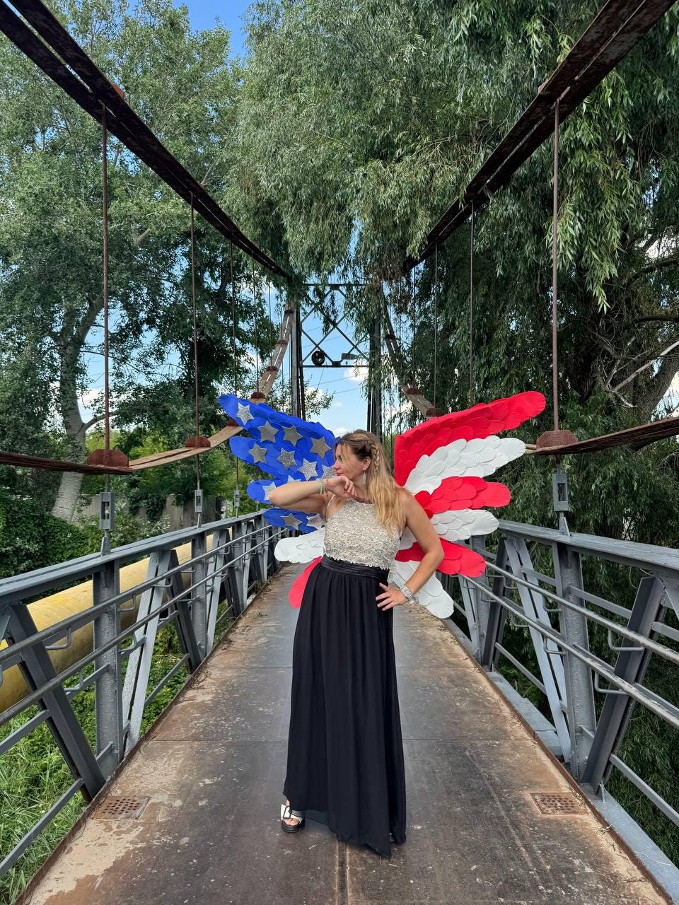 May include: A woman wearing a silver sequined top and a black maxi skirt stands on a bridge. She is wearing red, white, and blue wings, resembling the American flag. The bridge has metal railings and is surrounded by trees.