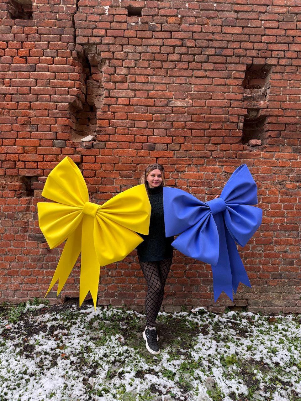 May include: A woman holding two large decorative bows. One bow is bright yellow, the other is a vibrant blue. The bows are against a weathered brick wall. The bows have long, flowing tails.