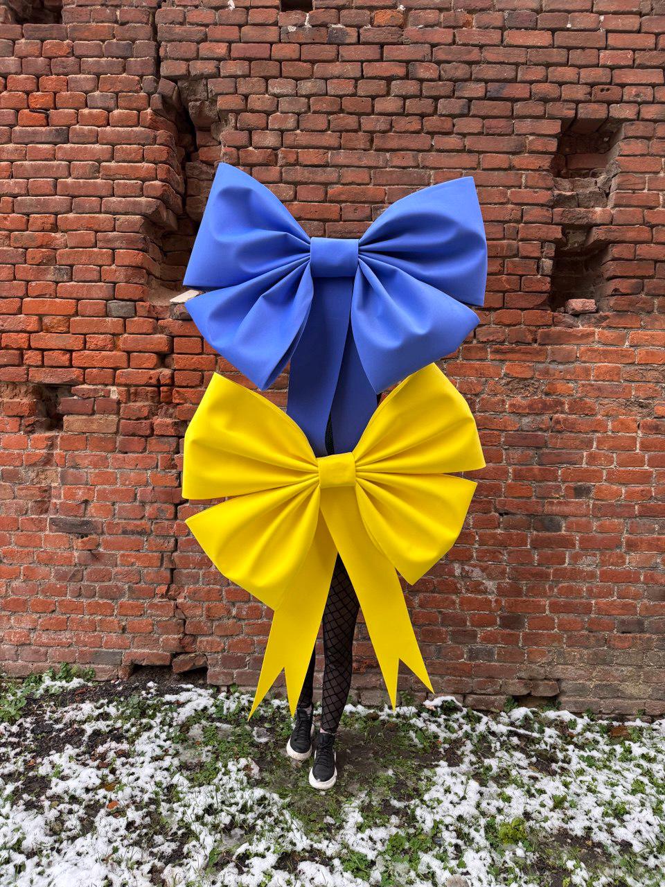 May include: A large blue bow is positioned above a large yellow bow. The bows are in front of a red brick wall. The bows are made of a stiff material. The person wearing the bows is wearing black shoes and black tights.