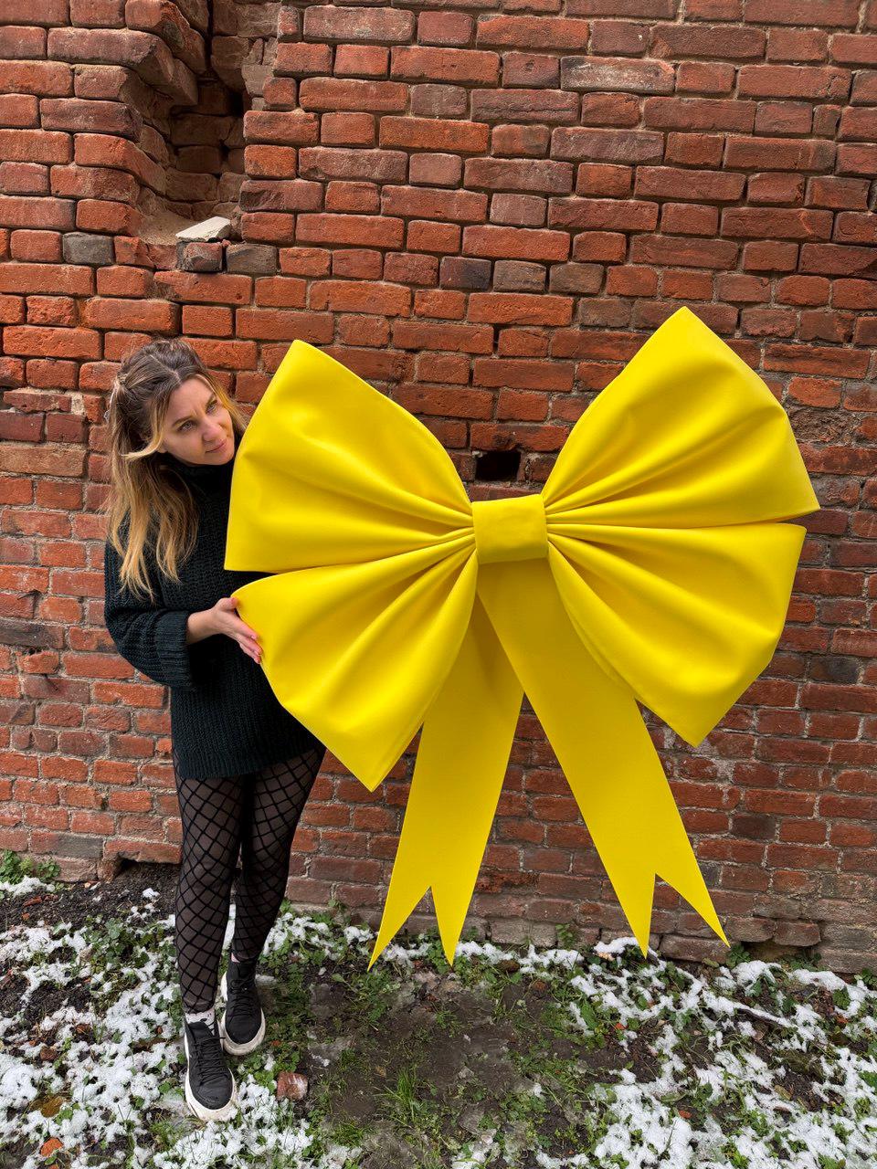 May include: A large, bright yellow bow with long, flowing ribbons. The bow is made of a smooth, possibly satin-like material. The background is a weathered brick wall.