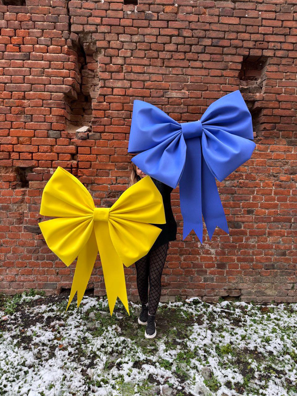 May include: Two large decorative bows, one yellow and one blue, are displayed against a weathered brick wall. The bows have long, flowing tails and are made of a smooth, matte material. The yellow bow is on the left, and the blue bow is on the right.