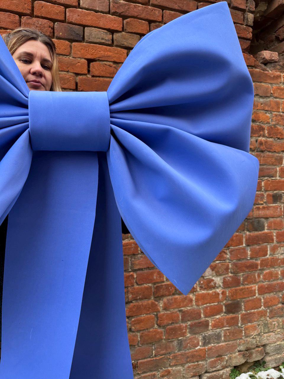 May include: A large, vibrant blue bow with a central knot and flowing tails. The bow is made of a smooth, matte material. The background is a weathered brick wall.